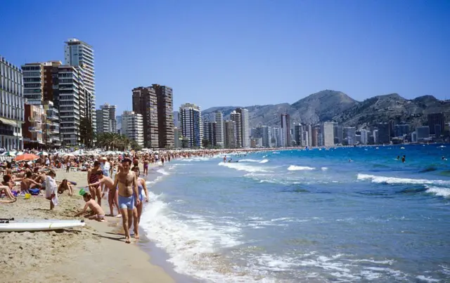 Vista de Benidorm desde la playa, con turistas bañándose y rascacielos. 