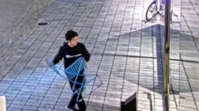 Мужчина с черными волосами в черном спортивном костюме поднимает синий складной стул, идя по улице к стеклянной двери
