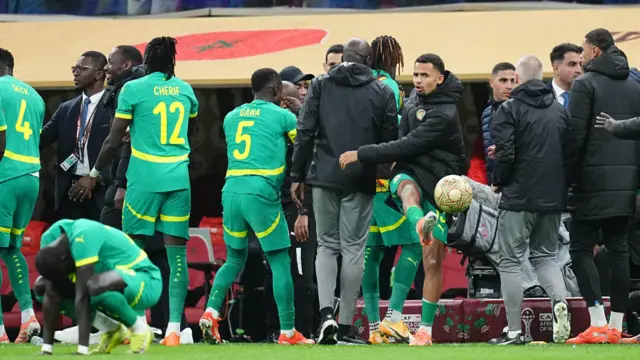Les joueurs et les entraîneurs sénégalais se tiennent sur la ligne de touche, la plupart dos à la caméra, lors de la finale de la CAN 2025 alors qu'ils protestaient contre la décision d'un arbitre. Iliman Ndiaye fait face à la caméra et est vu à mi-chemin en train de lancer un ballon sur le terrain avec colère.