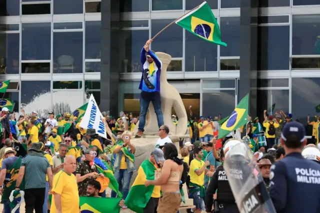 Centenas de pessoas em volta de estátua da Justiça em frente ao STF; há um manifestante em pé, em cima da estátua, erguendo bandeira do Brasil