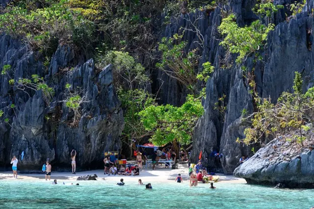 Pequeña playa entre rocas empinadas y algunos árboles, con varias personas nadando y en la arena, así como una mesa y venta de algo que no se alcanza a ver