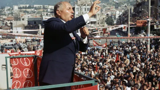 Milli Görüş hareketinin kurucusu Necmettin Erbakan, 1980 yılında Milli Nizam Partisi'nin bir mitinginde konuşuyor. Kürsünün çevresinde partinin logosu var. Önünde ise yüzlerce kişilik bir kalabalık görülüyor.
