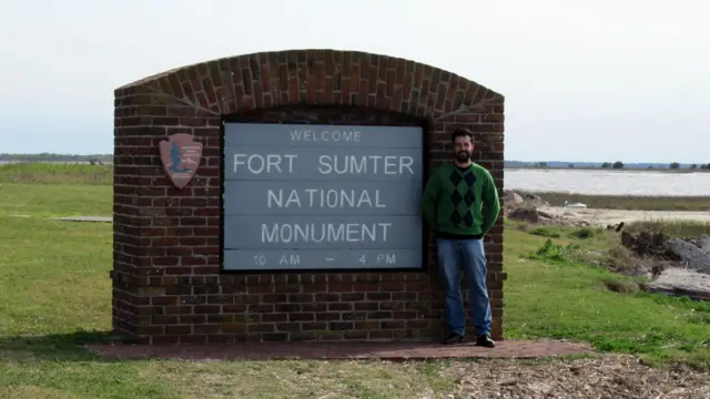 Célio Alcantara Silva posa ao lado de uma placa de tijolos do Fort Sumter National Monument, nos Estados Unidos. Ele veste um suéter verde com padrão em losangos escuros e jeans, enquanto sorri para a câmera. Ao fundo, aparece uma paisagem costeira aberta, com vegetação baixa, água e céu claro.