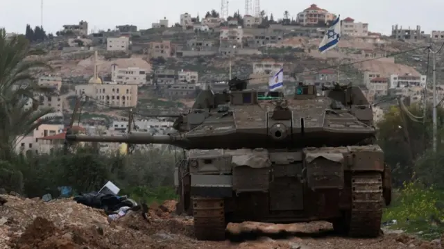 دبابة إسرائيلية ترفع علمي إسرائيل، وخلفها مبانً وبجانبها أشجار