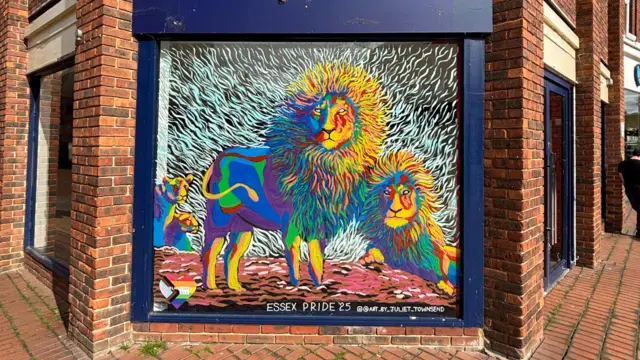Se han pintado leones arcoíris de colores en una ventana de una calle principal. El marco de la ventana es azul marino y los ladrillos que la rodean son marrones.