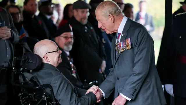 Король Карл в сером костюме с медалями на лацкане держит за руку мужчину в инвалидном кресле