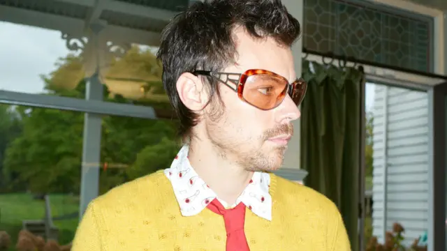 Harry Styles w żółtym swetrze, jaskrawoczerwonym krawacie i pomarańczowych okularach przeciwsłonecznych