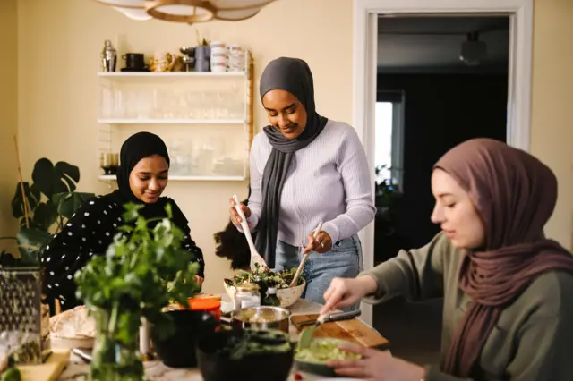 Femme souriante préparant une salade tandis que des amis préparent le repas à table dans la cuisine. 