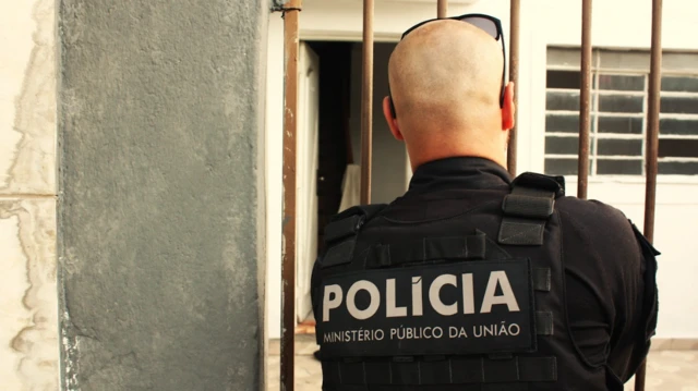 Homem com farda que fiz: Polícia - Ministério Público da União; ele está de costas para a foto e próximo a portão de casa