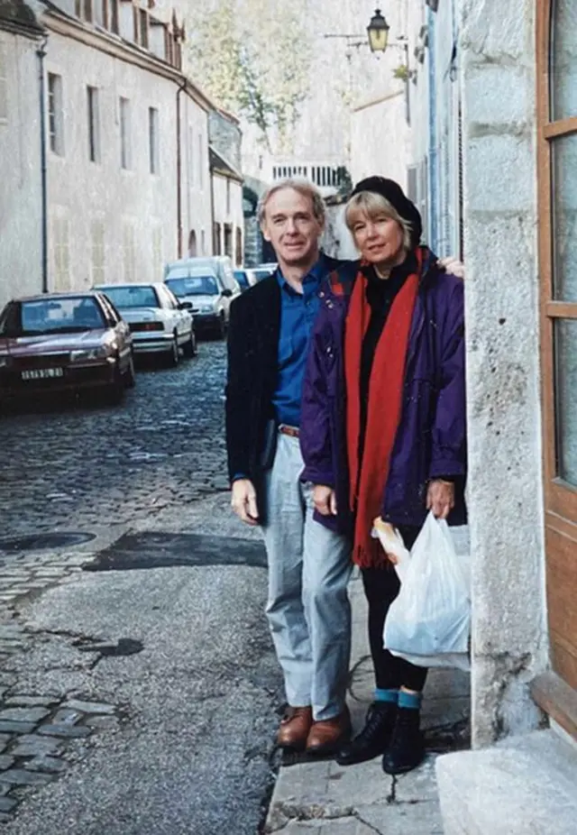 Chuck y Daphne posan en Beaune, Francia. Él viste unos vaqueros y una chaqueta de traje negra sobre una camiseta azul oscuro. Ella, quien lleva un abrigo morado, una bufanda roja y un gorro negro, sujeta una bolsa de plástico blanca.