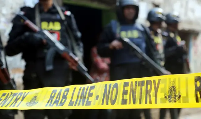 Yellow police tape that reads 'RAB line - no entry' is seen in front of members of the Rapid Action Battalion (RAB) standing guard outside a militant hideout in Dhaka on 12 January 2018. They stand carrying rifles with 'RAB' in yellow letters on their uniform and wear helmets.
