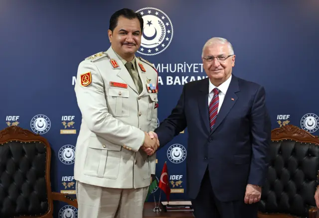 Saddam Hafter ve Yaşar Güler gülümseyerek ve tokalaşarak poz veriyor. Güler takım elbiseli, Hafter ise üniformalı.