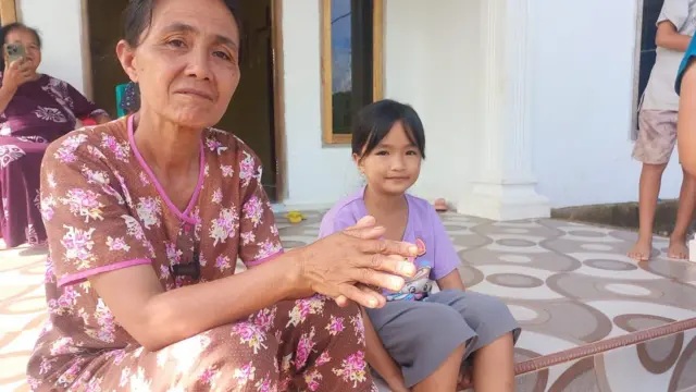 Yania Tilase menggunakan pakaian tidur motif bunga bersama cucunya sedang duduk di teras rumah, sambil melihat ke arah kamera.