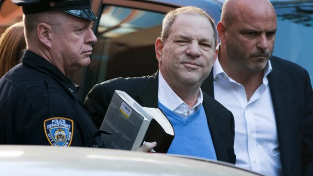 هاروی واینستین با دو کتاب در دست در میان یک پلیس و یک لباس شخصی