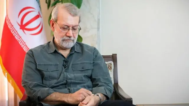 علی لاریجانی به ساعت مچی‌اش نگاه می‌کند و پرچم جمهوری اسلامی پشت سر اوست