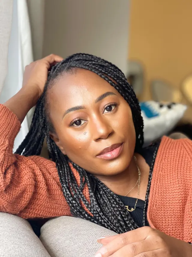 Destiny wear brown sweater, She put her hand for head as she dey look di camera.