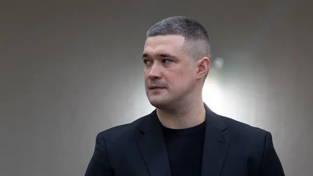 Портретне фото чоловіка в темному піджаку та чорній футболці на нейтральному розмитому тлі. Людина з короткою стрижкою дивиться вбік, знімок зроблено в приміщенні під час офіційного заходу або публічного виступу.