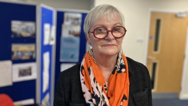 Julie Reader-Sullivan, directora ejecutiva de Headway Gloucestershire, mira a la cámara. Se encuentra en un edificio de oficinas; el fondo no está enfocado. Tiene el pelo corto y lleva gafas redondas, una bufanda estampada y una chaqueta oscura.