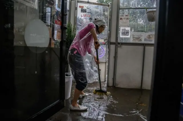 Mulher tentar retirar, com vassoura, água e sedimentos que entraram na casa