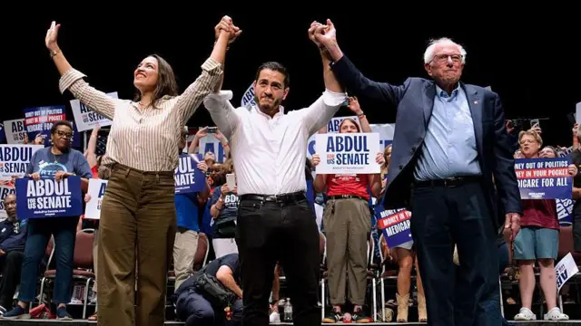 Alexandria Ocasio-Cortez, Abdul El-Sayed y Bernie Sanders están tomados de las manos y las levantan durante un evento de campaña en el que varias personas levantan carteles de Abdul para el Senado.