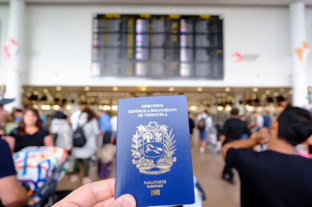 Un pasaporte venezolano delante de un panel que indica destinos de viaje en una terminal de pasajeros.