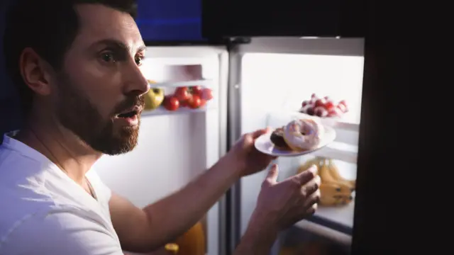 Homem branco e de barba vestido com camiseta branca segura prato de doces com porta de geladeira aberta durante a noite