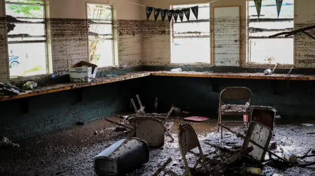 Imagem de uma sala com cadeiras jogadas cheia de lama.