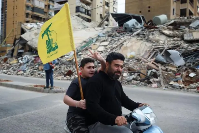 شخصان يستقلّان دراجة نارية صغيرة، يرفع أحدهما علم حزب الله ويشير بإصبعين في الهواء، في شارع في بيروت أمام كومة كبيرة من الركام حيث كان مبنى قائماً، في صورة التُقطت يوم الجمعة.