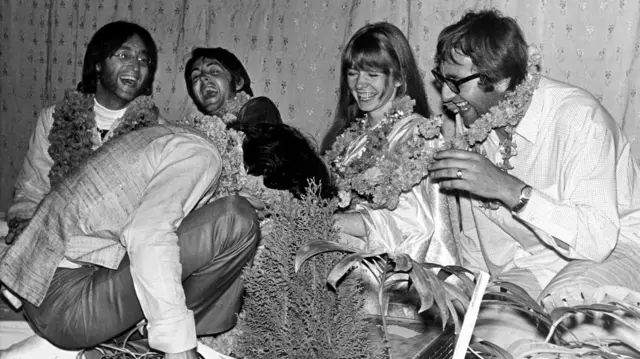 Учасники Бітлз і їхні друзі сидять на підлозі з квітковими гирляндами на шиях і сміються. 