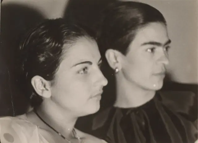 Perfil del rostro de Cristina, que tiene una blusa blanca y un collar. A su lado, de fondo, está Frida, que tiene una blusa negra y aretes. También está de perfil.