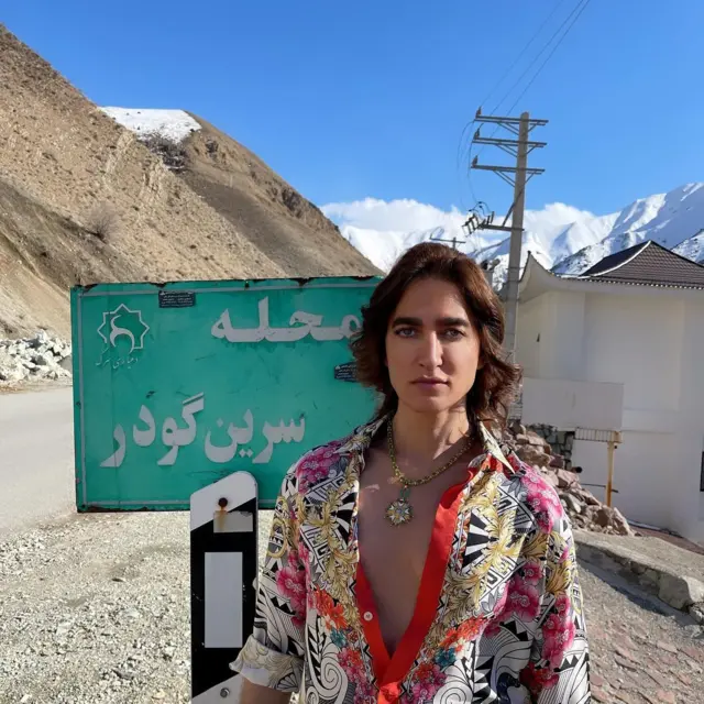 سرین بدیعی جلوی یک تابلوی شهرداری ایستاده است. روی تابلو نوشته است محله سرین گودر؛ در پس زمینه کوه‌های برفی دیده می‌شود. یک ساختمان و تیر چراق برق هم در سمت راست تصویر پشت سرین بدیعی قرار دارد