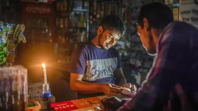 একটি দোকানে মোমবাতি জ্বালিয়ে কাজ করছেন একজন, সামনে আরেকজন দাঁড়িয়ে আছেন 