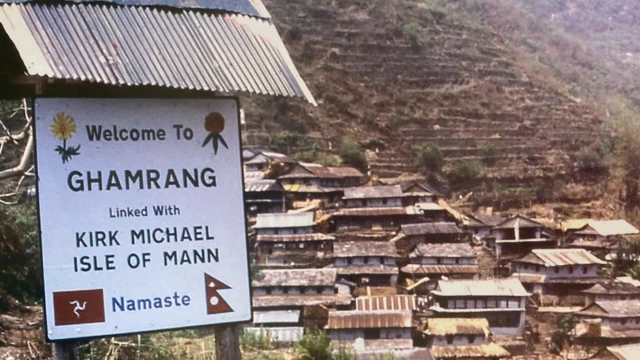 Imagen de un cartel en la región nepalí de Ghamrang. Indica que Ghamrang está conectado con Kirk Michael, Isla de Man. Al fondo se puede ver el pueblo circundante en la cima de la montaña.