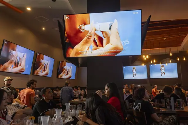 En el interior de un bar de deportes, se ven seis televisores sincronizados mostrando una publicidad de GLP-1.