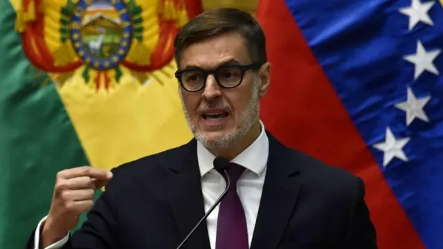 Félix Plasencia em primeiro plano, de terno e gravata, fala frente a um microfone com as bandeiras da Bolívia e da Venezuela ao fundo