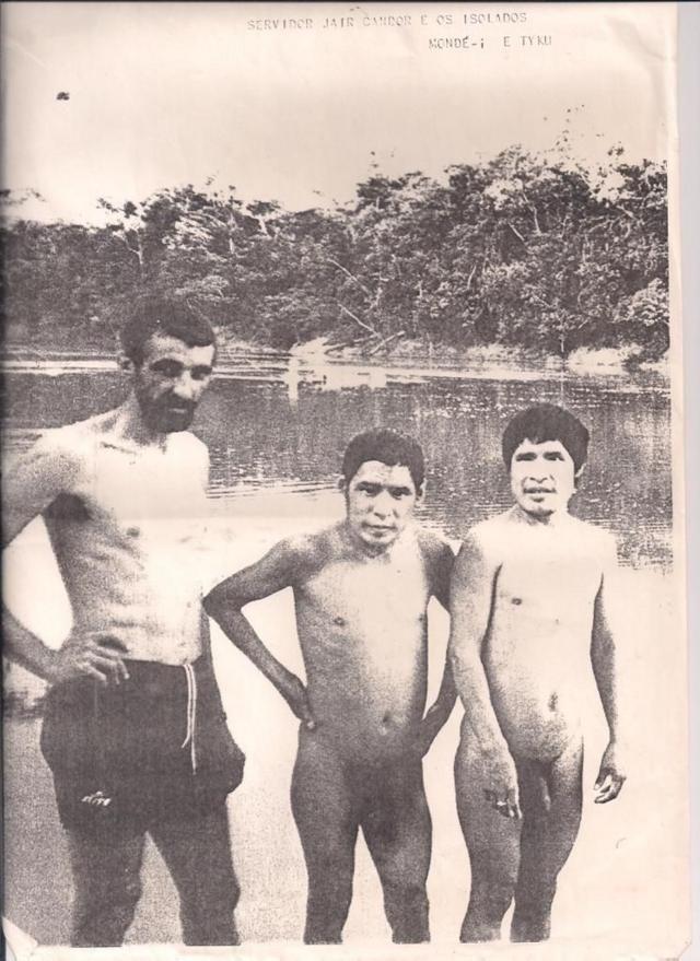Foto em preto e branco mostra homem branco ao lado de dois indígenas em frente ao rio