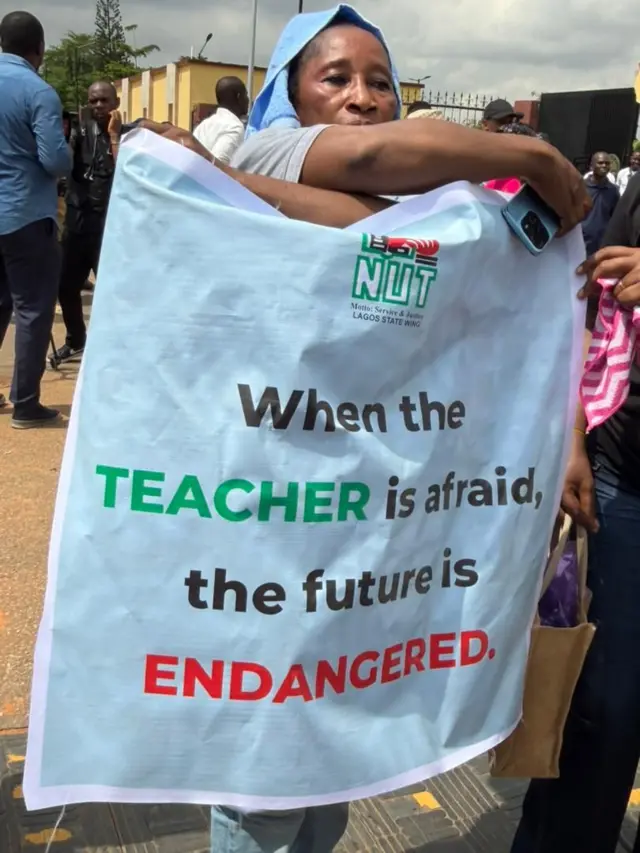 Sad looking woman dey hold banner wey say, "wen di teacjer dey afraid, di future dey in danger"