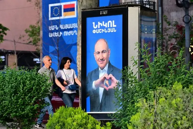 Paşinyan posterde, eliyle kalp emojisi yapmış, bir kadın ve erkek önünden geçiyor.