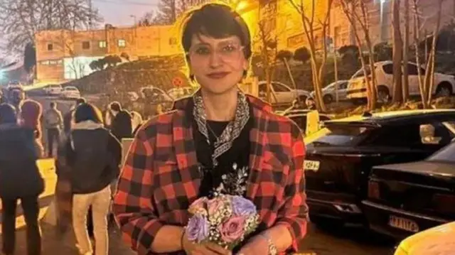 ফুল হাতে দাঁড়িয়ে আছেন মোতাহরেহ গুনেই, তার পর লাল ও কালো রংয়ের চেক শার্ট। পেছনে ডান পাশে কয়েকটি গাড়ি এবং বাম পাশে কিছু লোক দাঁড়িয়ে আছে