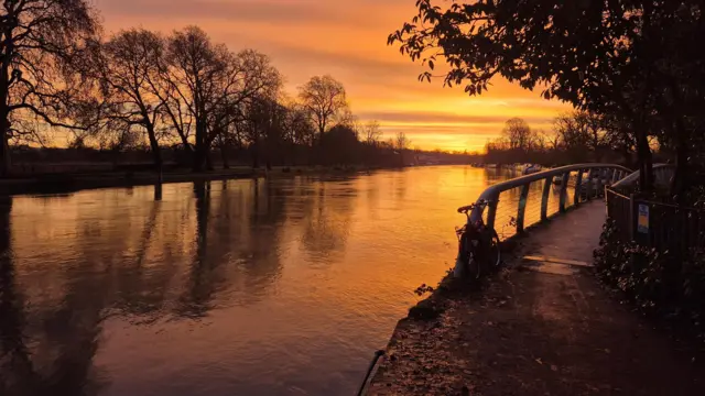 Un amanecer dorado sobre un río. El río está bordeado de árboles y hay un pequeño puente con una bicicleta apoyada. El cielo brilla de un naranja intenso y se refleja en el agua.