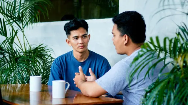 جلس رجلان ذوا شعر أسود عند طاولة خشبية بنية اللون، وعلى الطاولة كوبان أبيضان، ونباتات خضراء على جانبي الطاولة.