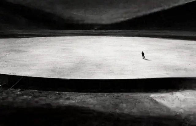 Черно-белая картинка, где посреди огромной овальной площадки видна фигура человека.