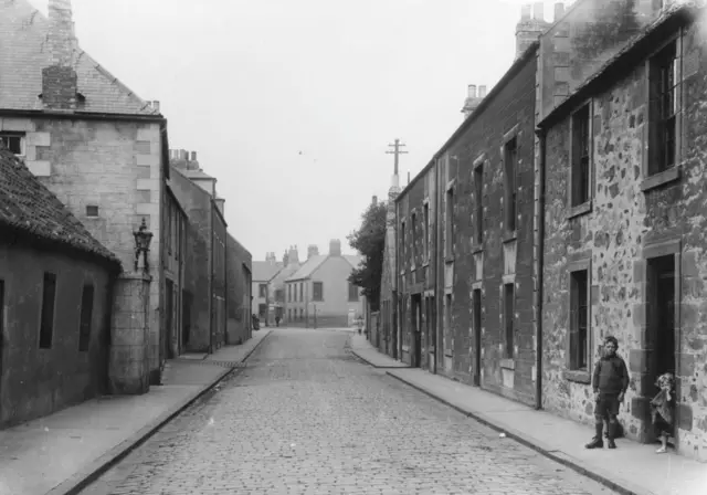 Uma rua de Eyemouth na década de 1930, em uma imagem em preto e branco, com duas crianças saindo de uma casa — não são Jim nem Betty.