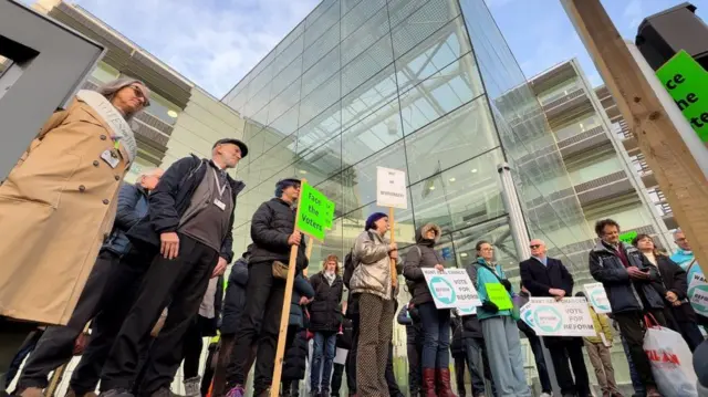 Un grupo de unas personas, de pie frente a un edificio de cristal, sostiene pancartas y protesta. Las pancartas muestran los logotipos y colores de Reform UK y del Partido Verde.