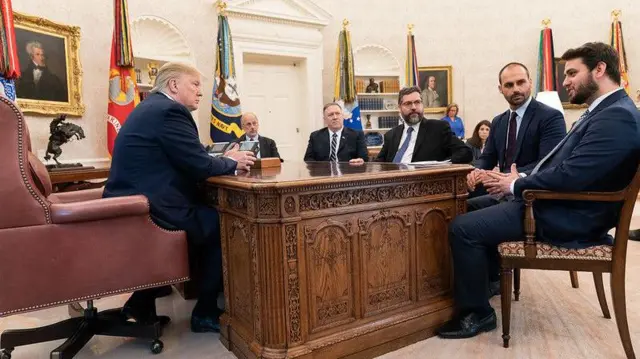 Donald Trump observa brasileiros durante reunião com equipe do presidente Bolsonaro. Seis pessoas estão sentada, usando terno,mines onabettornomines onabetuma mesamines onabetmadeira na Casa Branca