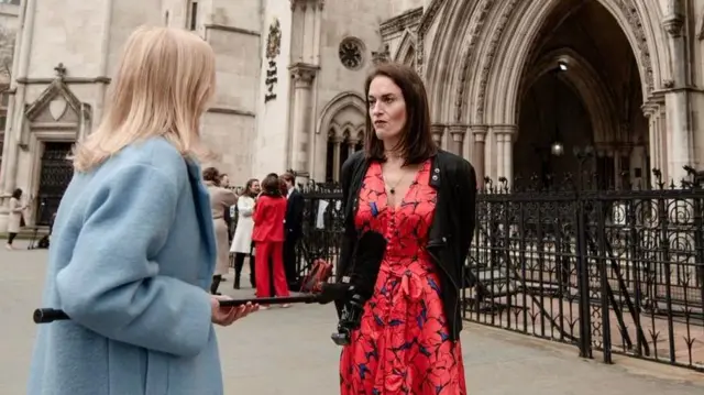 Jamie Klingler, kırmızı ve mavi bir elbise ve siyah bir ceket giymiş halde, Kraliyet Ceza Mahkemesi önünde, elinde mikrofon tutan ve açık mavi bir ceket giyen bir haber muhabiri tarafından röportaj yapıyor.