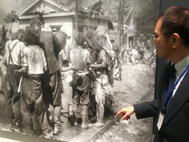 Мушкарац стоји поред фотографије деце која стоје у гомили после атомске бомбе у Хирошими