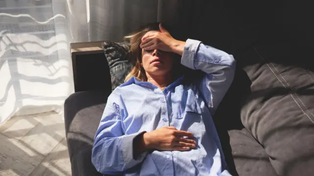 Mulher deitada em um sofá, aparentemente com dor de cabeça, ao lado de uma janela por onde entra forte luz do dia