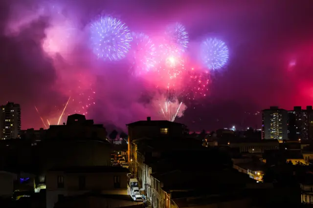 Una panorámica de edificios en la oscuridad con el cielo iluminado por fuegos artificiales azules y rojos
