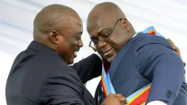 Joseph Kabila (ibumoso) na Félix Tshisekedi (iburyo) barimo guhererekanya ubutegetsi muri Mutarama (1) mu 2019. Bambaye amakositimu, barimo kumwenyura, Kabila yambika Tshisekedi igitambaro kinyuze mu kwaha no mu ijosi, gifite amabara y'ibendera rya DRC. 